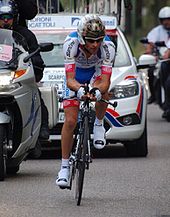 A solo cyclist competing in a time trial, being followed by a motorcyclist and a team car