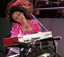 A mid shot of a female performer wearing bright pink clothes and playing a musical keyboard. Musical equipment and staging sits around her.