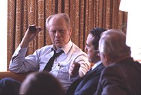 Photograph of Gerald Ford, seated with a pipe in hand, talking to three men