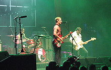 Photo from right side of stage shows Gillies behind the drum kit. Johns in right profile, singing into a microphone and playing a guitar. Joannou is playing his bass guitar with knees partly bent and looking at his left hand on the fret board. Equipment obscure the front of the stage.