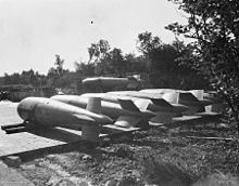 Black and white photograph of six aerial bombs resting on stands in an open area.