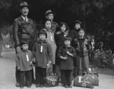 Dorothea Lange's photo of the Mochida family waiting for the bus in Hayward (1942). Hiroko and Miyuki are in the front row, at left; Miyuki is holding a sandwich. The children are wearing tags with the family number to help keep them together.