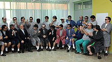 Color image of President Piñera visiting the rescued miners, who are dressed in hospital robes and pajamas while seated and standing in a semi-circle in a hospital room