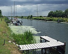 A broad, flat artificial cut with dark water and bright green raised banks. A patch of green algae stains the water. Several modern yachts are moored on wooden jetties spaced along the bank