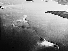 Black and white aerial photo of two ships sailing together near the shoreline. One of the ships is partially obscured by a large splash.