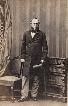 Sir John Duckworth standing between a chair to the left and low bookcase to the right, holding a top hat in his left hand