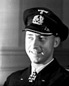 A smiling man wearing a peaked cap and dark military uniform with an Iron Cross displayed at the front of his uniform collar.