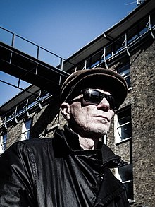 A 52-year-old man is shown in upper body shot, from below and to his right. He wears a brown corduroy cap, dark sunglasses and a black leather coat. His face is weathered and bristly. Behind him is a multi-storey building with a high walkway connected to an overhead footbridge.