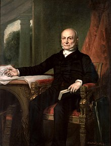 The official presidential portrait of John Quincy Adams by artist George Peter Alexander Healy. Adams, his head mostly bald and his face framed with a faint muttonchops beard, looks to the audience with firm gaze. He sits in an ornate chair, the arms decorated with sphinxes. In his left hand he holds a book, one finger inserted into the pages, perhaps to mark where he left off. His right hand gestures to a table next to him, where a sprawling sheet of paper is spread. No text is visible on the paper, but there is a portrait of George Washington, from the bust up, peeking from behind Adams's hand.