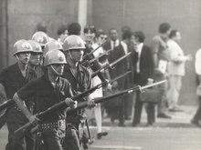 Several men in a line wear helmets and brandish rifles with bayonets
