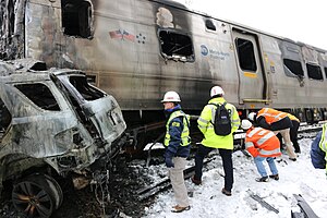 Several people in neon colored safety clothing and white helmets examine the fire-damaged wreckage of a silvery passenger rail car along the top with "Metro-North Railroad" written on it. One person has the letters "NTSB" on their jacket sleeve. At left is the rear of a similarly fire damaged automobile.