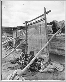 Black and white photo of women weaving on traditional loom, sheep in background