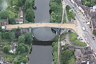 An aerial photo of the bridge, showing the close proximity of the main road alongside the river and the many building dotted nearby.