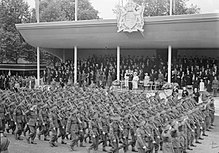 Black and white photo of a large number of men wearing military uniforms marching in close formation past an open-air stage