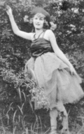 A black and white photograph of a young woman outdoors in a ballet pose, one arm extended. Her shoulders are bare, and she is wearing a wide-brimmed hat. She is looking directly at the camera.