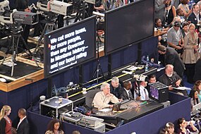 (B) The fourth teleprompter: A large confidence monitor displays the scrolling text of the speech immediately below the lenses of the broadcast TV cameras, several meters/feet away from the speaker.