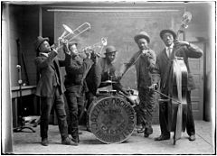 A black-and-white photo of a jazz band – consisting of two trombonists, a drummer, a double bassist, and a violinist – dressed in suits and bowler hats.