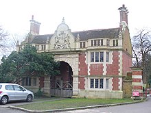 A photograph of a three-story brick building with a prominent archway in the centre surmounted by an elaborate carved crest. The sides of the building are topped by a set of triple chimney-stacks.