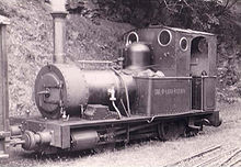 A small steam locomotive stands beside a banked area. On the left hand coal bunker is painted TAL-Y-LLYN RAILWAY. The track is covered with grass, and the rails are barely visible. In the foreground are some disused rails.