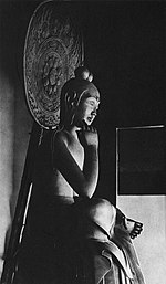 Three-quarter view of a seated statue in half-lotus position. The right foot rests on the left upper leg, the right elbow rests on the right knee with the right hand close to the head. The hair of the statue is sculpted with two top-knots. There is a halo behind the statue's head.