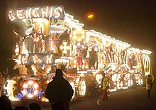 Night time photograph of lorry and trailer illuminated with thousands of light bulbs to make pictures. In the foreground and to the side are pedestrians.