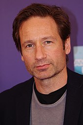 A man with dark brown hair is smirking and looking near the camera.