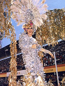 Rogéria standing on top of a platform with a dress covered in reflective silver feathers and a hat with peacock feathers.