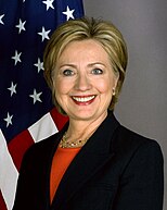 Hillary Clinton wearing a dark jacket over an orange blouse. The United States flag is in the background.