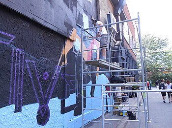 Young people standing on a ladder spray painting art on the side of a building