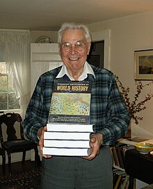 Smiling older man holding a stack of books in front of him; the top one is tilted up so the title, World History, is visible.