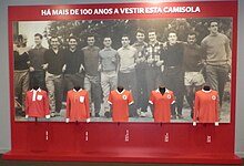 Five different Benfica shirts are displayed on a panel titled "HÁ MAIS DE 100 ANOS A VESTIR ESTA CAMISOLA" ("wearing this shirt for over 100 years") with a background picture of twelve Benfica footballers casually dressed and lined up.
