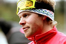Seitliches Porträt von Tomas Gustafson. Seine gelbe Skibrille sitzt auf der Stirn, darunter ein buntes Stirnband. Außerdem ist der Kragen einer roten Jacke oder eines roten Anzugs zu sehen.