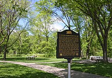 A park with several trees and picnic tables behind a plaque titled "A Golden Dedication for I-90"