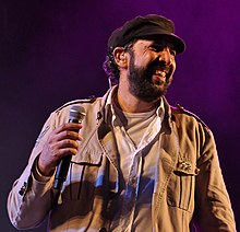 A bearded man with a black hat and a brown jacket is holding a microphone on his left hand and facing left