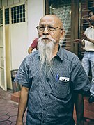 Mong Hay standing in a street. He is bald with a long white bear and wearing a collared, short sleeved shirt with a pen and smart phone in the pocket.