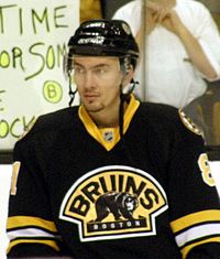 An ice hockey player standing, facing towards the camera. He is wearing a black helmet and a black, yellow and white uniform.