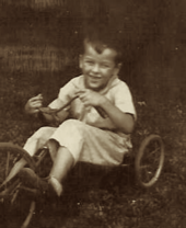 Toddler Ted Stevens rides on a tricycle; he has a smile. The image is in sepiatone.