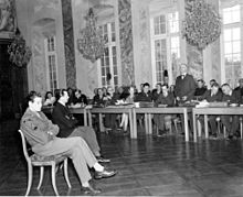 An American soldier interprets for a witness, a German, to the character of one of the defendants of the Borkum Island massacre. The witness is being addressed by a lawyer (standing, center right) of the defense counsel.