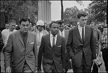 Chief U.S. Marshal James McShane and Assistant Attorney General for Civil Rights, John Doar of the Justice Department, are pictured escorting James Meredith to class at Ole Miss after the riot. Large groups of federal agents and likely students are seen in the background.