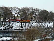 A freight train pulled by a locomotive is seen in the distance, across a moderately sized river. It is winter, and there is snow on the ground.