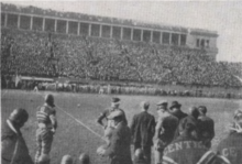 A group of football players standing on the sideline of a football field before a large crowd seated in the stadium