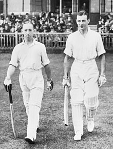 Two small men aged around 30 walk off the steps of a cricket ground pavilion onto the grass towards the centre of the playing area. Both wear light-coloured shirts, with rolled up sleeves, trousers and pads to protect their legs, all white. They wear batting gloves and hold a bat in readiness for play. The crowd sit behind a wooden fence; most are wearing suits and black cylindrical hats.