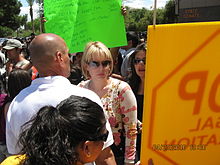 A woman in her thirties with fairly short blond hair, wearing sunglasses and a beige and pink top, is surrounded by a crowd.