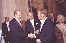 "Two men in formal wear, one the President of France and one President of the United States, greet a third man in formal wear during a state dinner party."