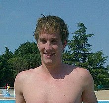 Sébastien Rouault steht mit nacktem Oberkörper und trockenen Haaren am Rande eines Schwimmbeckens, im Hintergrund sind grüne Bäume vor blauem Himmel zu sehen.