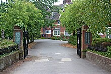 A gated entrance lined with trees leads to a red bricked building with white windows and a dark roof.