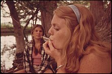 A young woman looking left with her eyes nearly closed smokes a large cigarette in a wooded setting near a body of water. Behind her another young woman looks at her