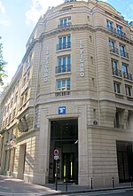 Frontale Farbfotografie eines hellbraunen, fünfgeschossigen Gebäudes mit dem Eingang an der Straßenecke. Über dem hohen Eingangstor ist das quadratische Logo mit dem weißen Buchstaben F auf hellblauem Untergrund. Links und rechts der oberen Fenster steht in weißer Schrift vertikal „Le Figaro“. Die Fenster haben französische Balkone.