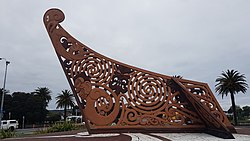 An elaborate Māori carving painted reddish brown carved like the end of a waka canoe set into concrete