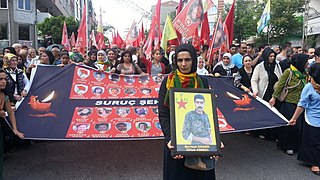 Porträt des YPG-„Märtyrers“ Şevket Demir (Çîya Amed) vor Transparent der „Suruç-Märtyrer“[63]